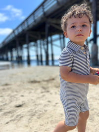 small boy with curly brown hair wearing our blue/cream stripe pique polo romper on the beach in summer.