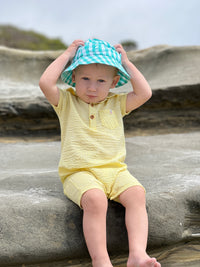 small blonde hair toddler wearing yellow seersucker polo and green plaid bucket hat, sitting on big rock at the beach in summer