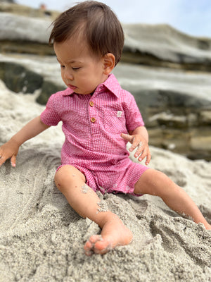 brown hair small boy wearing the coral seersucker polo romper sitting on rocks at the beach