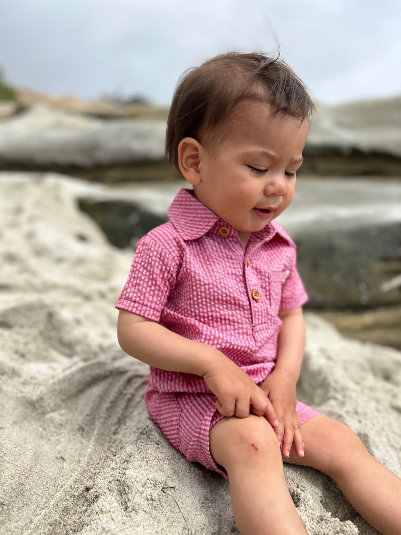 brown hair small boy wearing the coral seersucker polo romper sitting on rocks at the beach