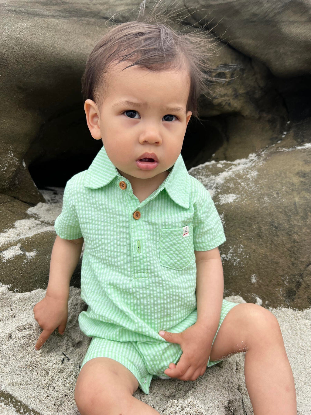 brown hair small boy wearing the green seersucker polo romper sitting on rocks at the beach