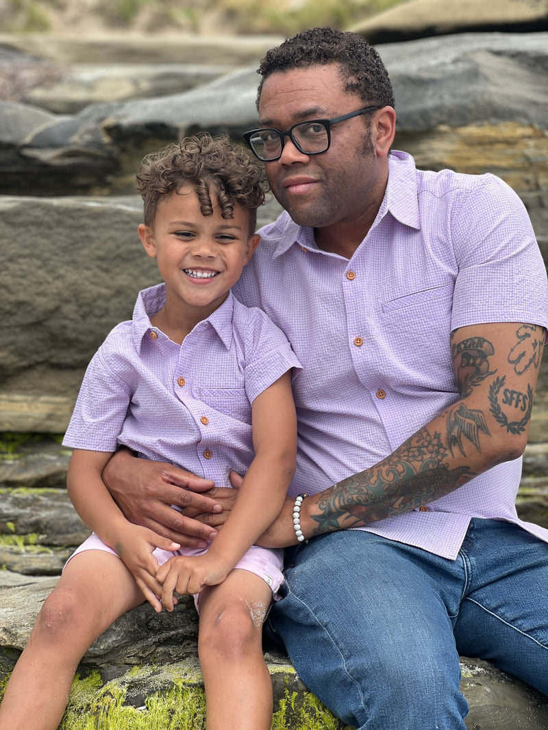 A boy with brown curly hair wearing our pink/lilac seersucker woven shirt and pink twill shorts, sitting on a big rock with his dad who has curly brown hair and is wearing the matching shirt in dad sizes.