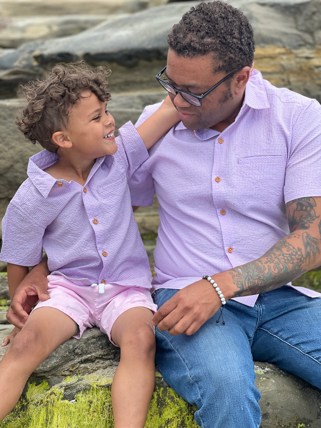 A boy with brown curly hair wearing our pink/lilac seersucker woven shirt and pink twill shorts, sitting on a big rock with his dad who has curly brown hair and is wearing the matching shirt in dad sizes. 