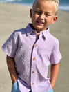 Blonde hair boy wearing our blue/pink seersucker wove shirt and blue pique shorts at the beach, smiling.