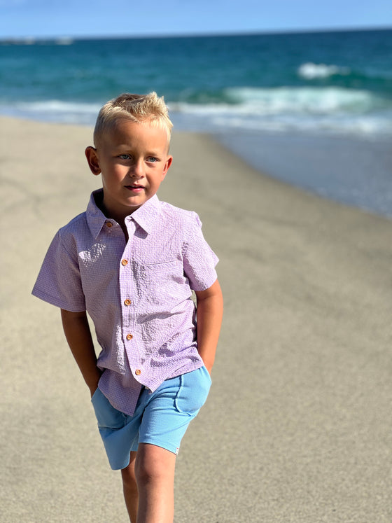 Blonde hair boy wearing our blue/pink seersucker wove shirt and blue pique shorts at the beach.