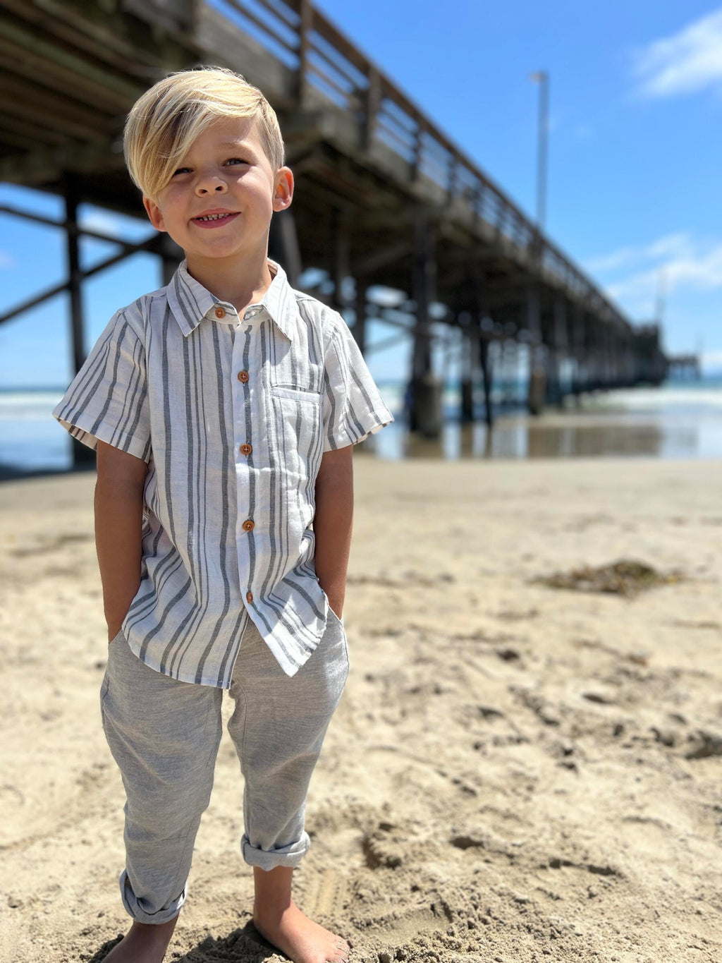 Blonde hair boy wearing our blue/white stripe woven shirt and grey gauze pants, standing on the beach.