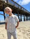 Blonde hair boy wearing our blue/white stripe woven shirt and grey gauze pants, standing on the beach.