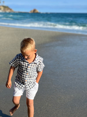 blonde hair boy wearing navy/white plaid woven shirt with white crew shorts, walking on the sand at the beach in summer