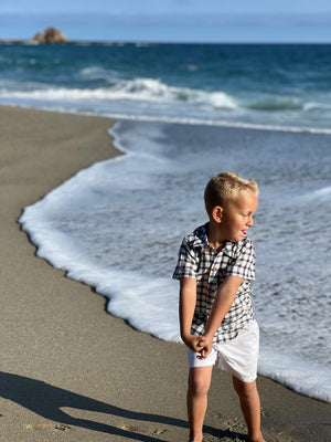 blonde hair boy wearing navy/white plaid woven shirt with white crew shorts, walking on the sand at the beach in summer