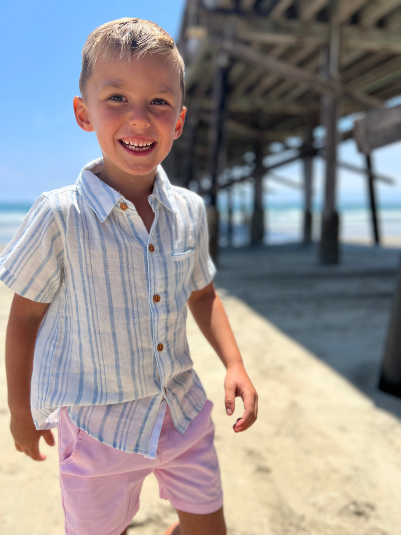 blonde hair boy wearing our blue/white stripe woven shirt and pink twill shorts walking on the beach.