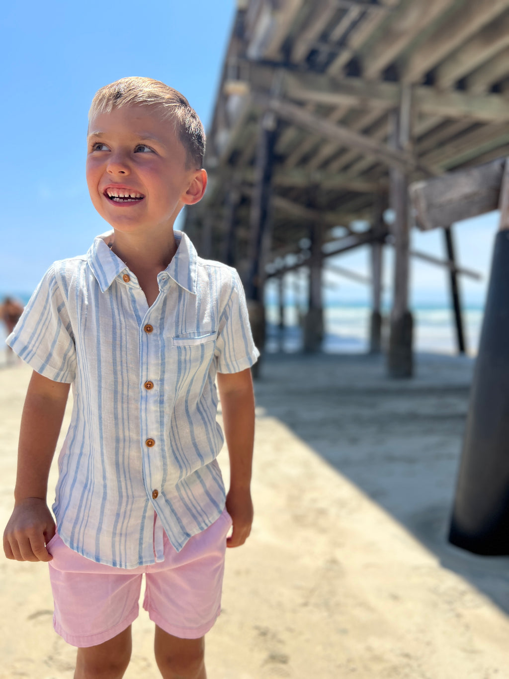 blonde hair boy wearing our blue/white stripe woven shirt and pink twill shorts walking on the beach.