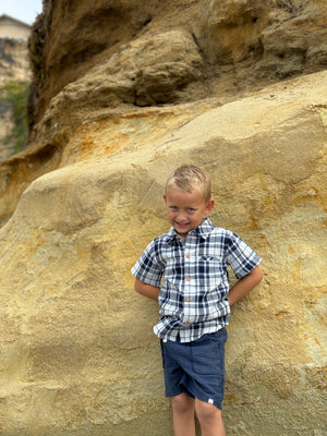 Blonde hair boy, with blue eyes wearing the navy/white plaid woven shirt and navy twill shorts posing in front of large beach rock