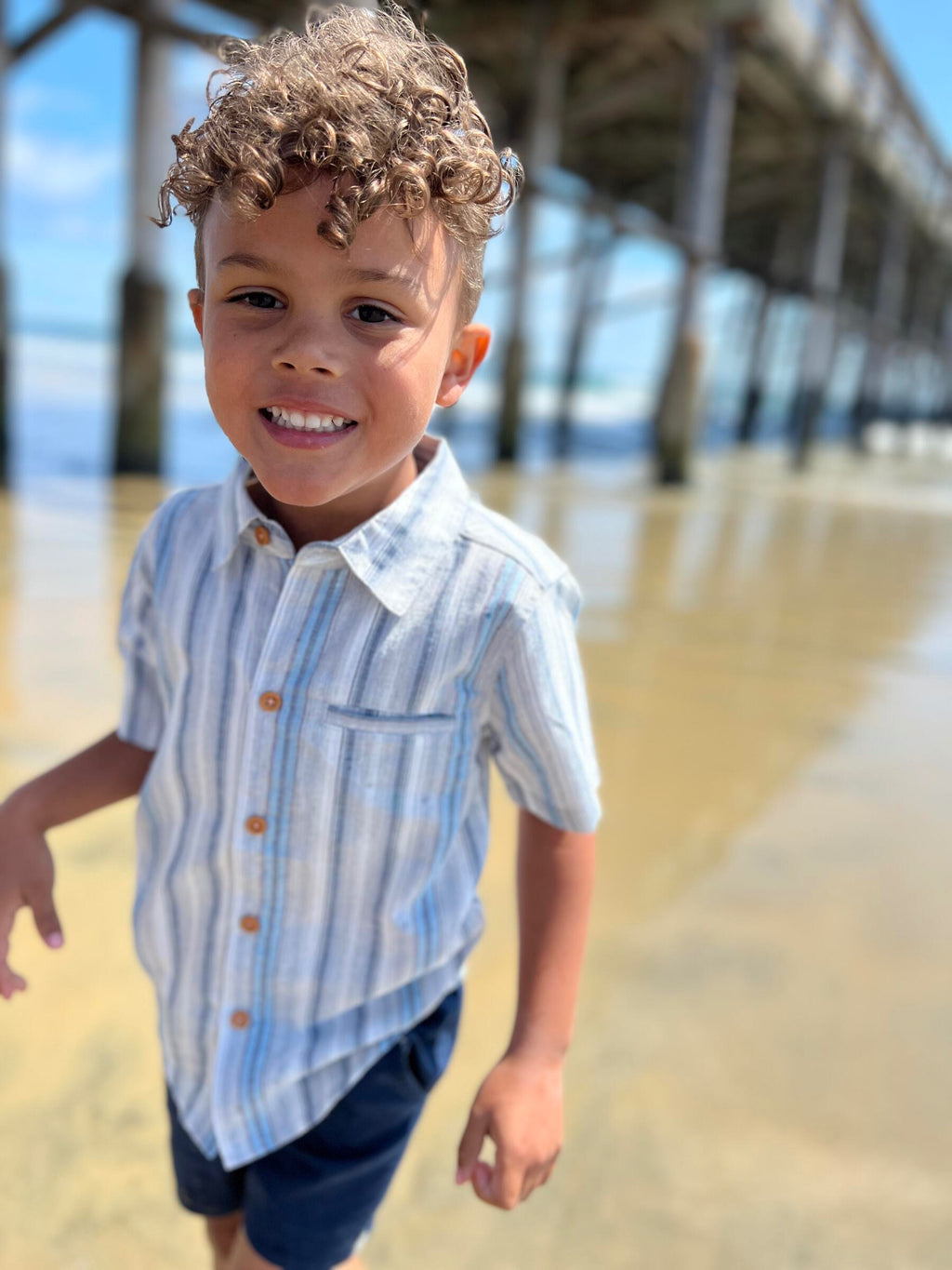 Brown curly hair boy wearing our blue stripe woven shirt and navy twill shorts walking on the beach.