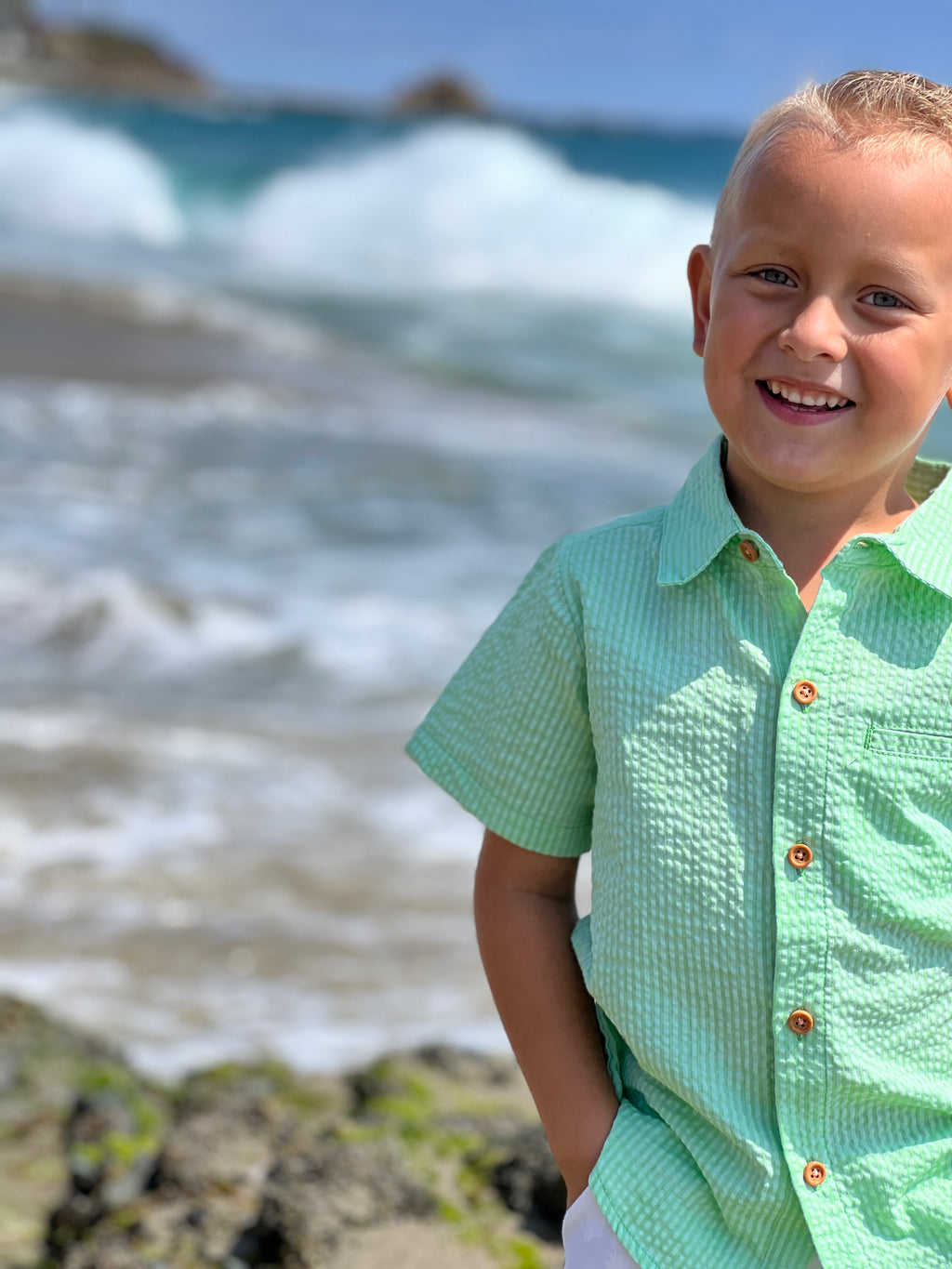 blonde hair boy with blue eyes, wearing Lime Seersucker Woven Shirt , standing at the beach, smiling at the camera.