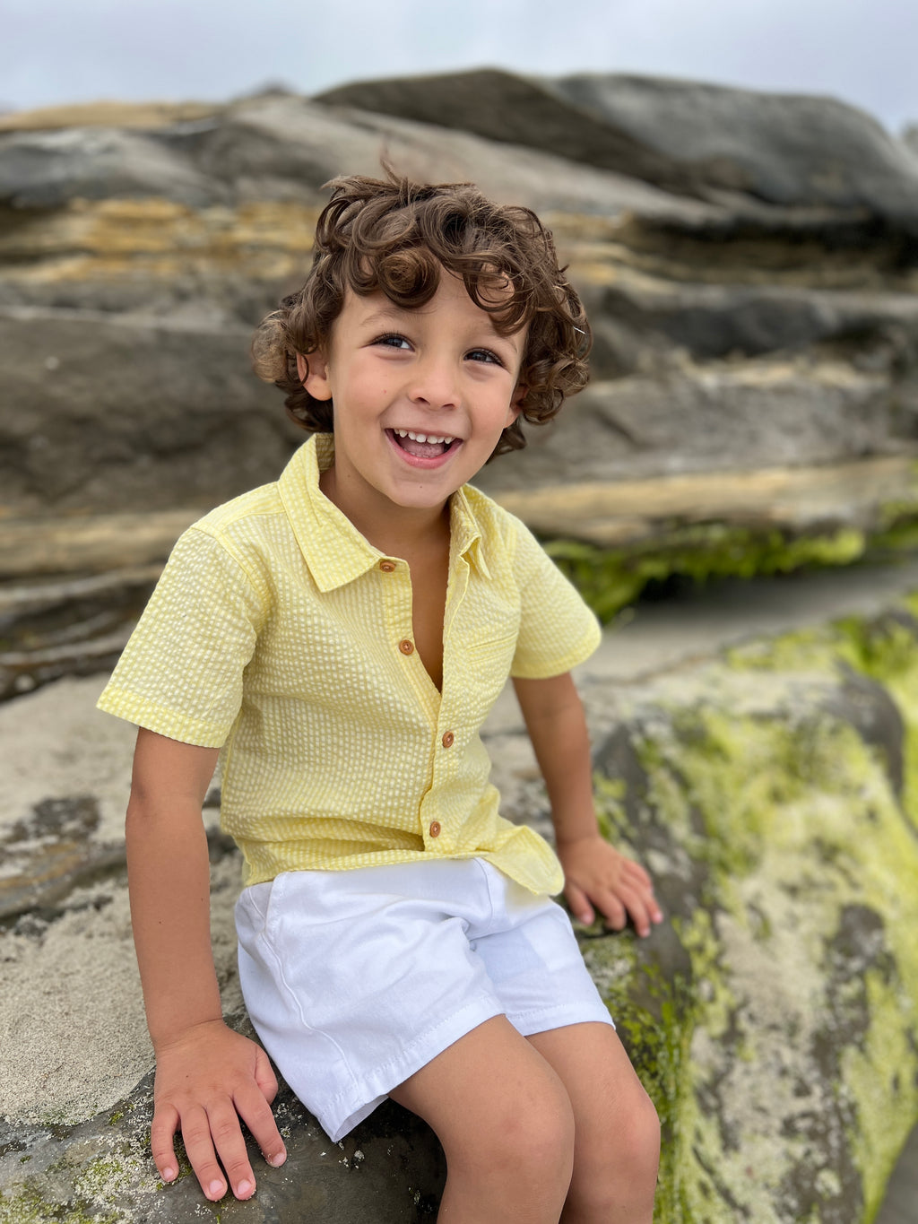 boy with brown curly hair and brown eyes, wearing our Seersucker Woven Shirt, and white gauze shorts sitting on a big rock at the beach