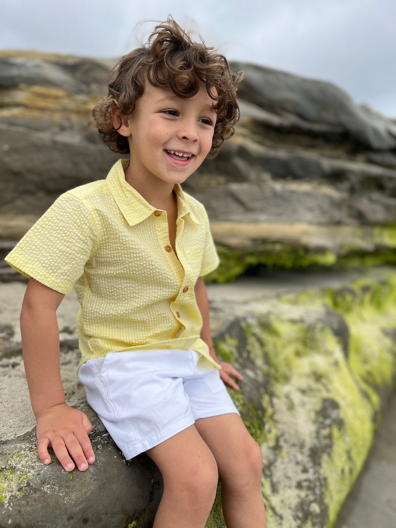boy with brown curly hair and brown eyes, wearing our Seersucker Woven Shirt, and white gauze shorts sitting on a big rock at the beach