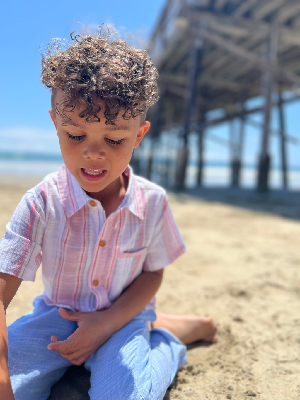 Small boy with curly brown hair, wearing our red/white/blue stripe woven shirt and blue heather pants sitting on the beach playing in the sand.