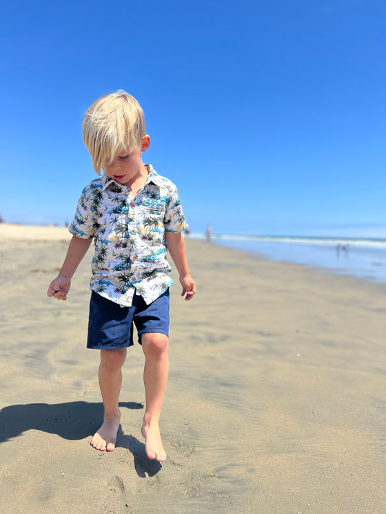 blonde hair boy wearing the cream Hawaiian print woven shirt and navy twill shorts walking along the sand at the beach.