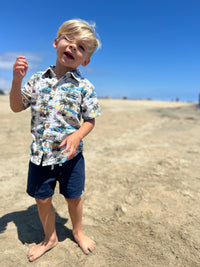 blonde hair boy wearing the cream Hawaiian print woven shirt and navy twill shorts walking along the sand at the beach.