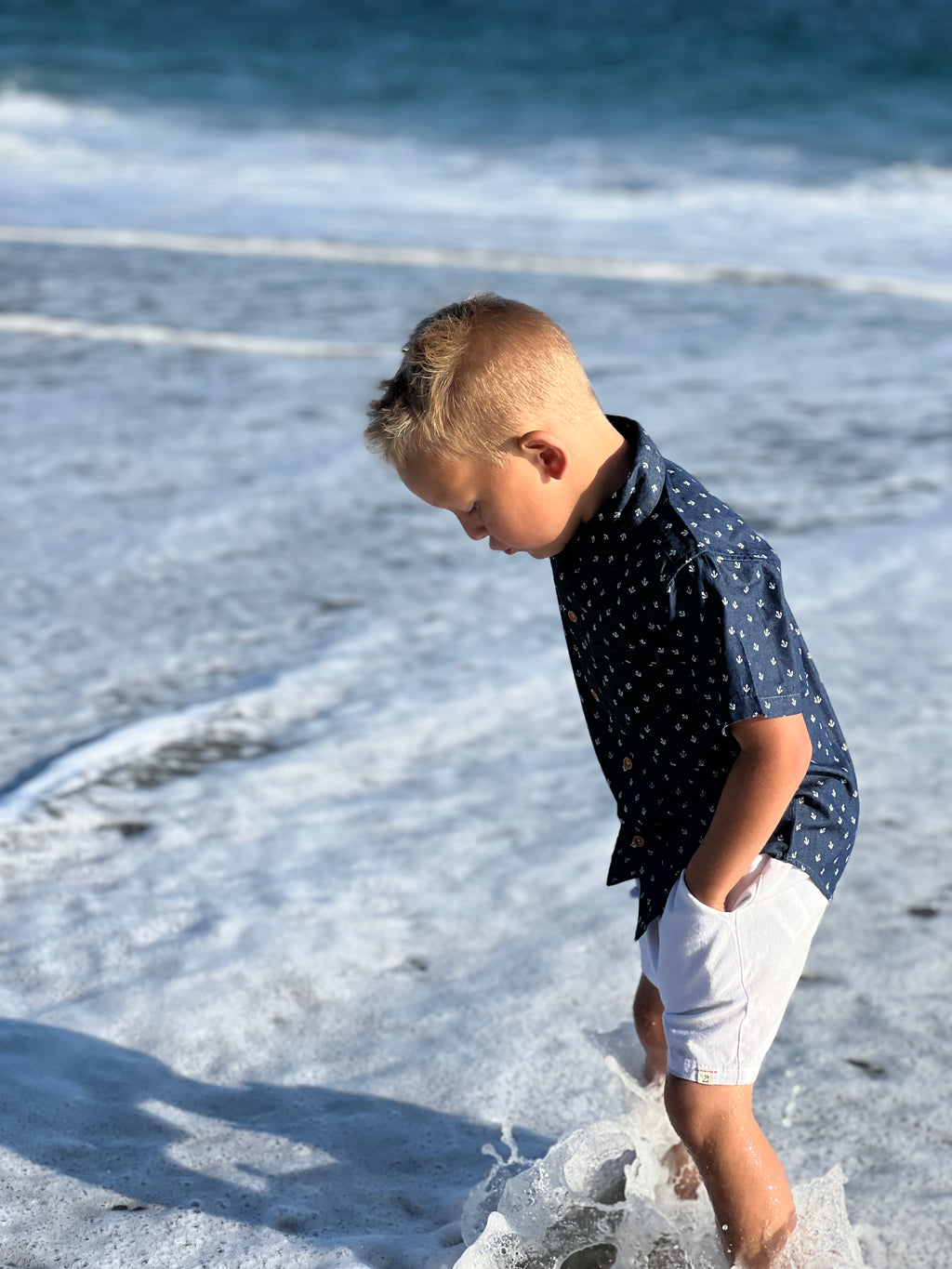 blonde hair boy wearing the chambray anchor woven shirt and white gauze shorts, splashing in the sea at the beach in summer.