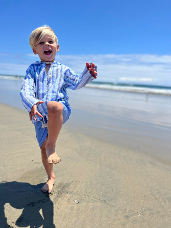 small blonde hair boy wearing a navy/white hooded top and pale blue pique shorts, jumping in the sand on the beach in summer