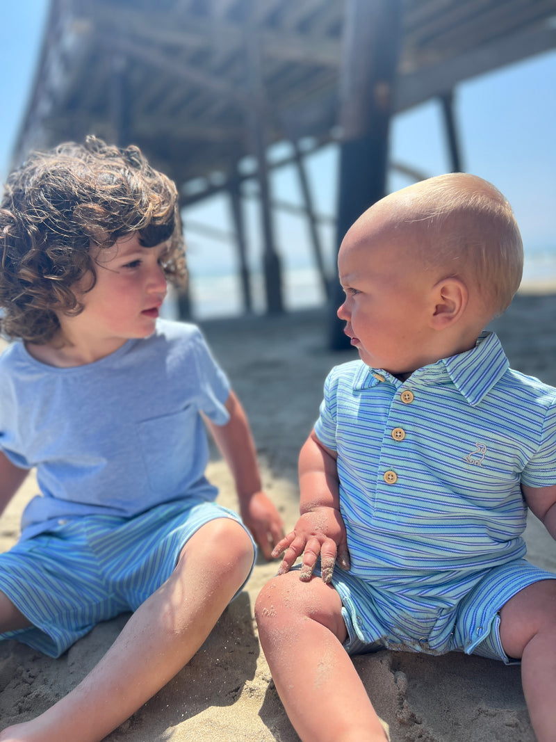 one brown curly haired boy wearing a blue tee with aqua blue stripe pique shorts and a blonde hair baby boy wearing an aqua blue stripe pique polo romper sitting on the sand at the beach in summer.