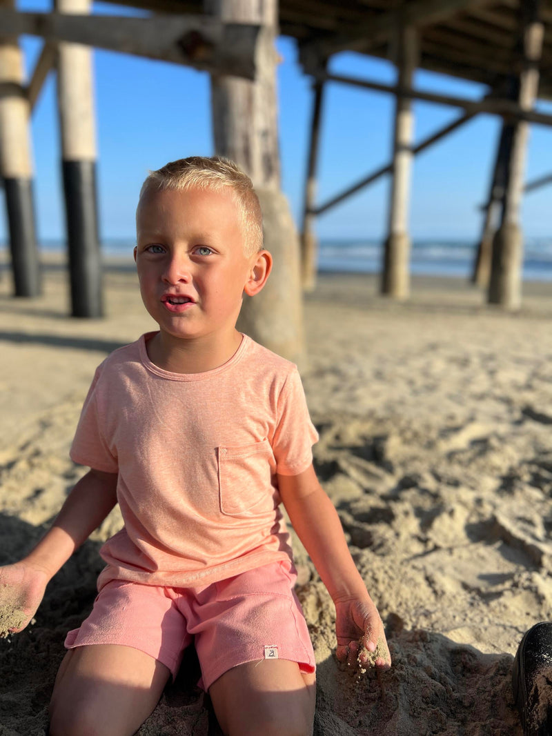 Blonde hair, blue eyed boy wearing peach tee and pink pique shorts playing in the sand at the beach in summer