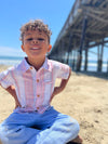 Small boy with curly brown hair, wearing our red/white/blue stripe woven shirt and blue heather pants sitting on the beach playing in the sand.