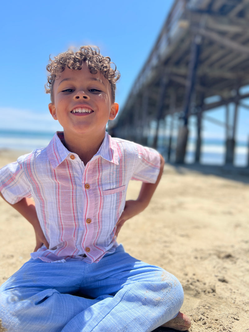 Small boy with curly brown hair, wearing our red/white/blue stripe woven shirt and blue heather pants sitting on the beach playing in the sand.
