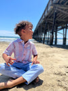 Small boy with curly brown hair, wearing our red/white/blue stripe woven shirt and blue heather pants sitting on the beach playing in the sand.