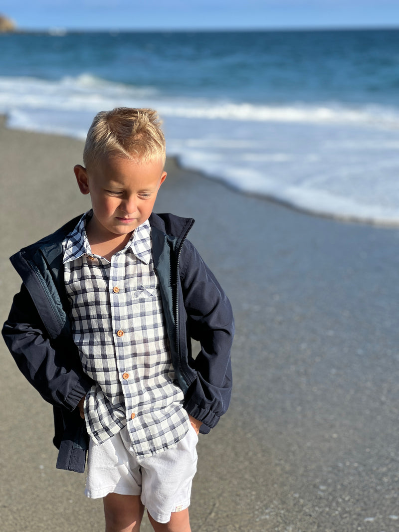 blonde hair boy wearing navy/white plaid woven shirt and the navy cloudbuster hooded lightweight jacket with white crew shorts, walking on the sand at the beach in summer