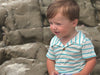 Child wearing a Turquoise striped Jersey onesie and White shorts sitting on rocks.