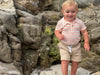 Child in Peach/White striped Jersey onesie Beige shorts standing on a rocky beach