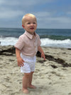 Child standing on a beach with ocean waves in the background ,Wearing a Peach Texture woven onesie and white shorts.