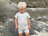 Fair haired boy standing on the sand in a henley grey ribbed romper