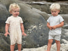 two boys playing on the sand wavey haired boy in a beige ribbed romper the other boy in a grey ribbed romper.