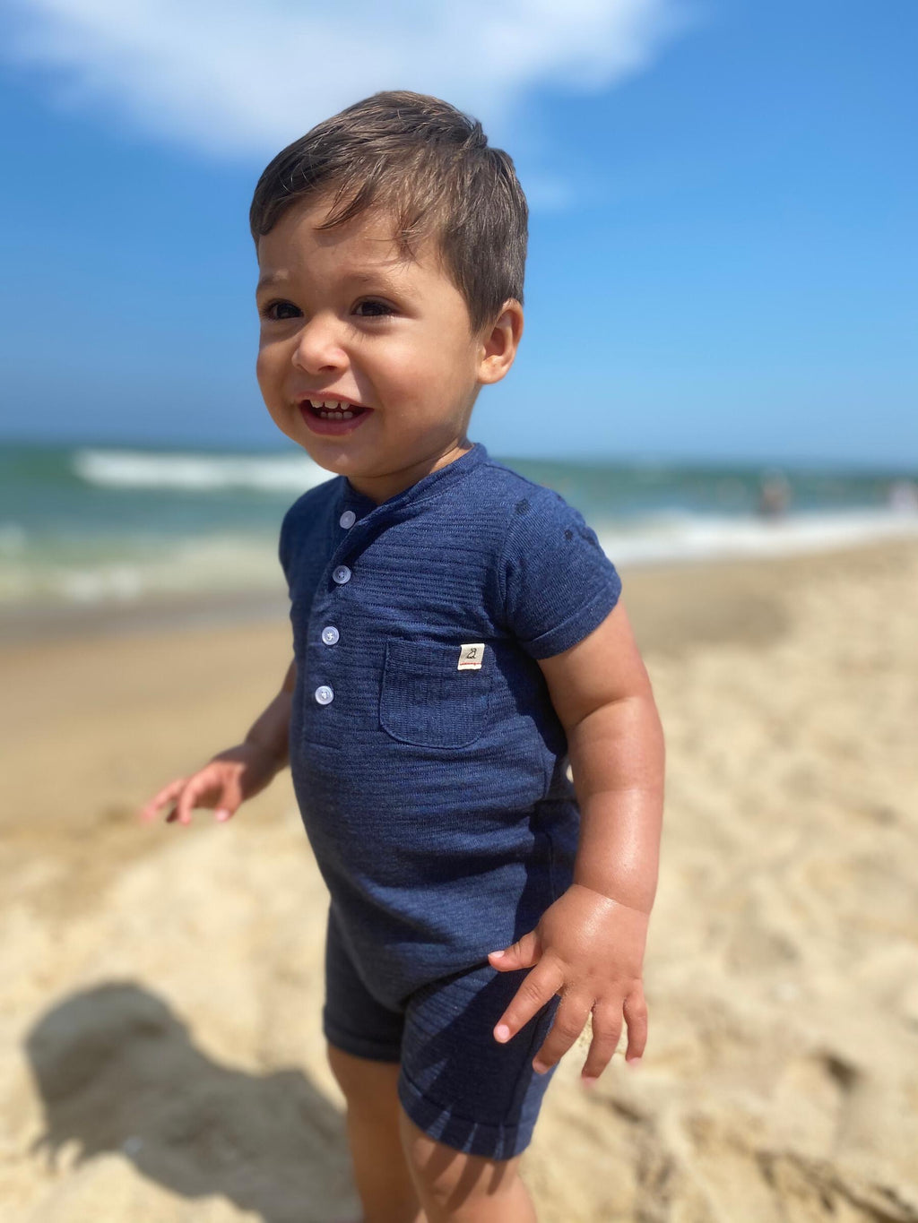 small boy with brown hair and brown eyes wearing the navy ribbed henley romper, on the beach in summer