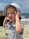 Child wearing a light grey bucket hat on a beach with grey woven onesie