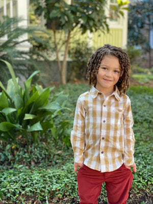 little boy, curly brown hair, wearing mustard/ white plaid woven shirt, red twill pants, in garden, long green grass, bushes and trees