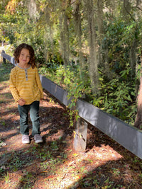 little boy, brown curly hair, wearing mustard henley stripe tee, fern trousers and grey Velcro shoes, walking in the woodlands