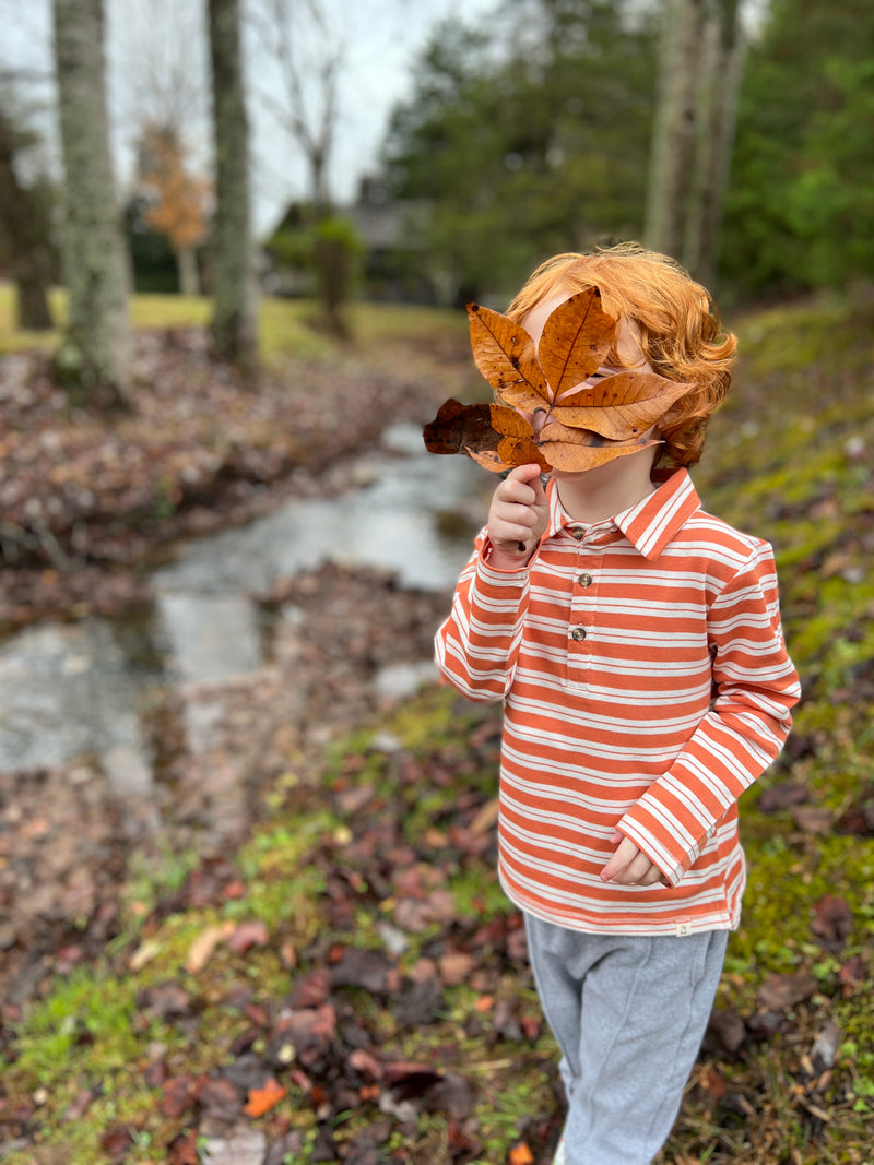 grey jog pants, white drawstrings, cuffed ankles, flexible waistband,cotton, polyester, spandex, little boy, ginger hair, pumpkin/white striped polo, holding autumn leaf, woodland, small stream, bright day, leaves on the floor, mud