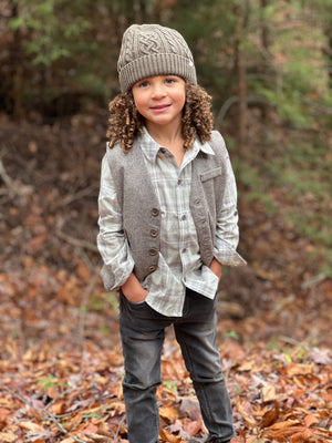 little boy, curly brown hair, grey knitted beanie, sage/ grey plaid woven shirt, grey tweed sleeveless vest, charcoal denim jeans, in the woodlands, autumn leaves on the floor
