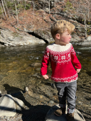 little boy , red cotton sweater, snowflake print and zig zag designs at the top and bottom of sweater, long sleeved, cuffed, wrists, cream collar, cream designs, fairisle, christmassy, cotton, soft, layer, with black denim jeans, red fairisle sweater, blonde boy, in woodland, stream, rocks,bright day, fern suede boots