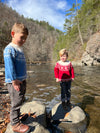 2 boys , Blue cotton sweater, snowflake print and zig zag designs at the top and bottom of sweater, long sleeved, cuffed, wrists, cream collar, cream designs, fairisle, christmassy, cotton, soft, layer, with black denim jeans, red fairisle sweater, blonde boys, in woodland, stream, rocks,bright day