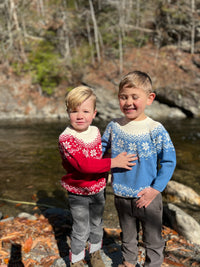 2 boys , Blue cotton sweater, snowflake print and zig zag designs at the top and bottom of sweater, long sleeved, cuffed, wrists, cream collar, cream designs, fairisle, christmassy, cotton, soft, layer, with black denim jeans, red fairisle sweater, blonde boys, in  woodland, stream, rocks,bright day