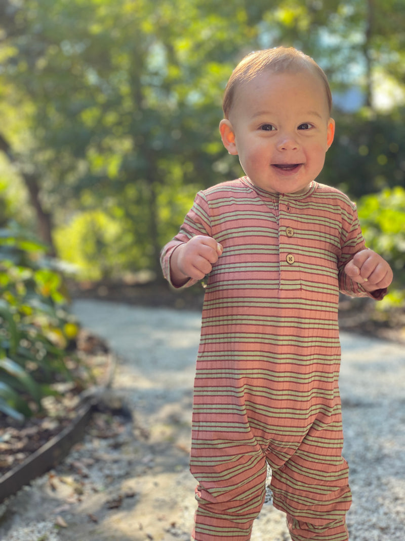little baby boy, brown hair, wearing dusty pink stripe romper, outside on pavement, trees in background