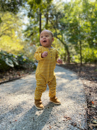 toddler, brown hair, wearing mustard stripe romper, outside on pavement, bushes in background, brown Velcro shoes