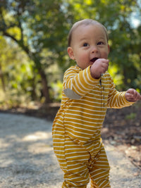 toddler, brown hair, wearing mustard stripe romper, outside on pavement, bushes in background