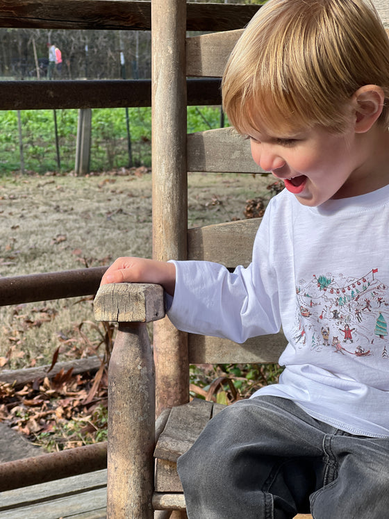 little blonde boy wearing white snow scene long sleeve, sleeved, sleeve, smart, casual , tee, charcoal denim jeans, sitting on wooden chair outside