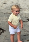 Child in a yellow striped jersey onesie and white shorts standing on a sandy beach.